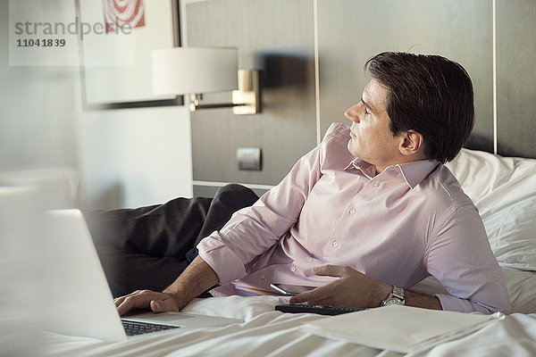 Geschäftsmann auf Hotelbett liegend mit Handy und Laptop-Computer  in Gedanken wegschauend