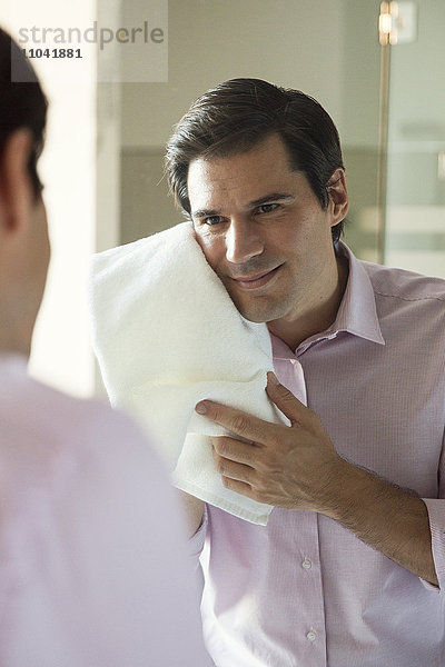 Mann schaut in den Spiegel und trocknet sein Gesicht mit einem Handtuch.