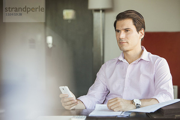 Geschäftsmann hält Smartphone im Büro  schaut in Gedanken weg