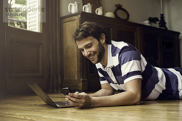 Mann auf dem Boden liegend mit Laptop und Smartphone