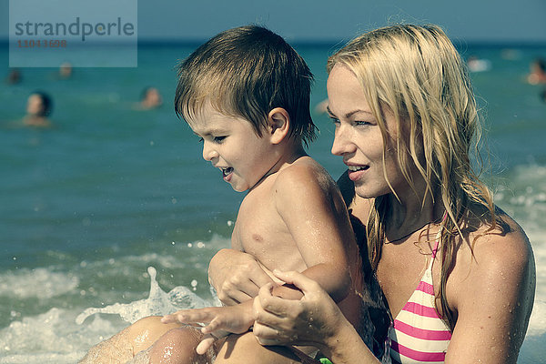 Eine Welle spritzt auf eine Mutter  die ihren Sohn am Strand hält