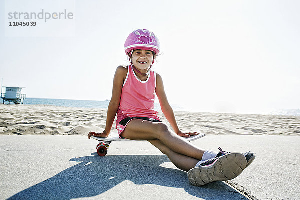 Gemischtrassiges Mädchen sitzt auf einem Skateboard am Strand