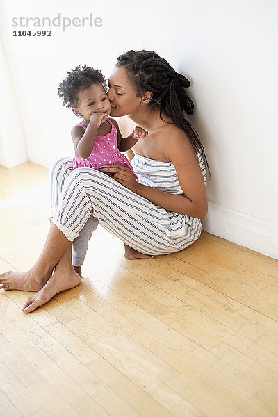 Schwarze Frau sitzt auf dem Boden und küsst die Wange ihrer kleinen Tochter
