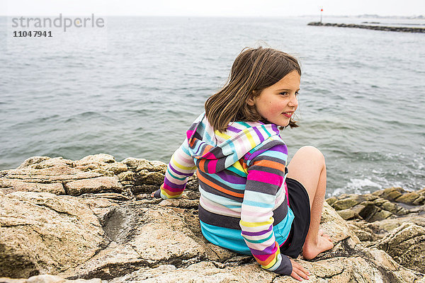 Lächelndes kaukasisches Mädchen sitzt auf Felsen am Meer