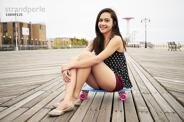 Gemischtrassige Frau sitzt auf einem Skateboard auf der Promenade