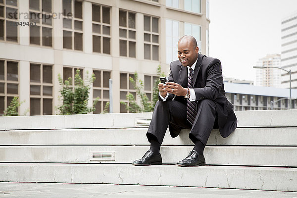 Schwarzer Geschäftsmann sitzt auf einer Treppe und schreibt eine SMS mit seinem Handy im Freien