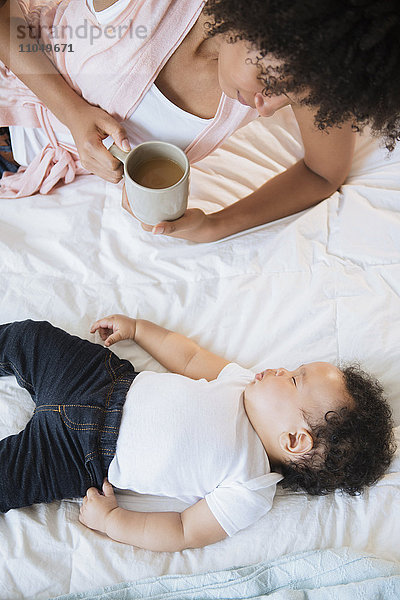 Mutter trinkt Kaffee  während sie ihren kleinen Sohn auf dem Bett schlafen sieht