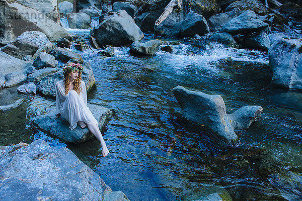 Kaukasische Frau mit Blumenkrone auf einem Felsen am Fluss