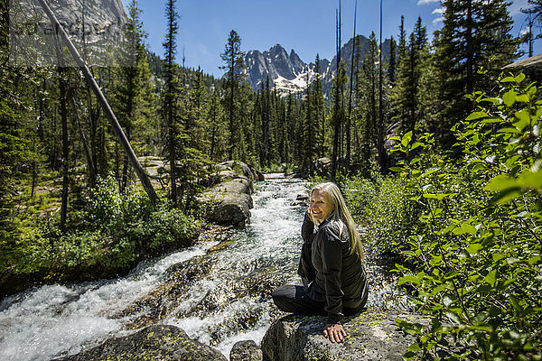 Lächelnde kaukasische Frau sitzt auf einem Felsen am Bergfluss
