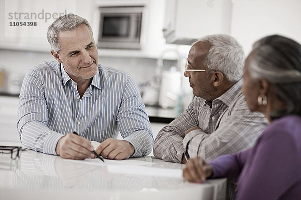 Drei Personen an einem Tisch  ein älteres Ehepaar sitzt mit einem Mann zusammen  der ihnen mit Papier und Stift Informationen gibt.