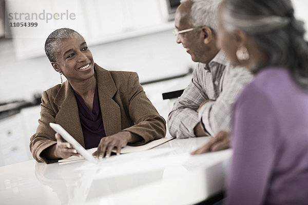 Drei Personen an einem Tisch  ein älteres Ehepaar sitzt zusammen mit einer Frau  die ihnen einen digitalen Tablet-Bildschirm zeigt.