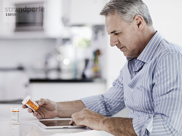 Ein Mann  der an einem Tisch sitzt  ein digitales Tablett benutzt  ein Tablettenfläschchen mit Medikamenten in der Hand hält und das Etikett liest.