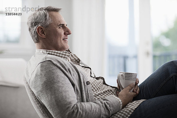 Ein reifer Mann mit grauem Haar  der sich auf einem Stuhl zurücklehnt und sich bei einer Tasse Tee oder Kaffee entspannt.