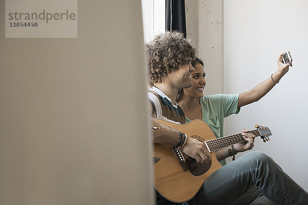 Loft-Wohnen. Ein junger Mann  der Gitarre spielt  und eine Frau neben ihm  die sich mit einem Smartphone ein Selfy nimmt.