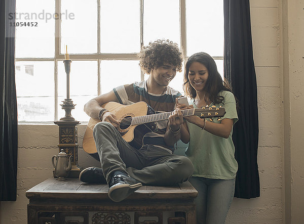 Loft-Wohnen. Ein junger Mann spielt Gitarre und neben ihm eine Frau  die mit einem Smartphone fotografiert.