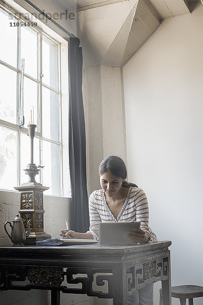 Loft-Wohnen. Eine Frau sitzt an einem Tisch am Fenster und benutzt ein digitales Tablett.