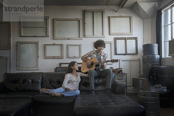 Loft-Dekor. Eine Wand  an der Bilder in Rahmen aufgehängt sind  die umgekehrt die Rückseiten zeigen. Ein Mann spielt Gitarre und eine Frau sitzt auf einem Sofa.