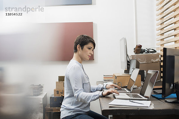 Eine Frau in einem Home-Office  die an einem Schreibtisch mit zwei Laptops sitzt  die Hände auf der Tastatur des einen Computers und den Blick auf den Bildschirm des anderen gerichtet.