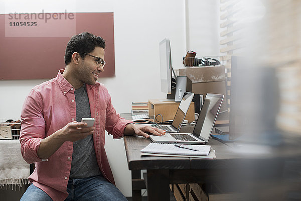Ein Mann in einem Heimbüro mit einem Schreibtisch mit zwei Laptops  der Informationen auf dem Bildschirm überprüft und ein Smartphone in der Hand hält.