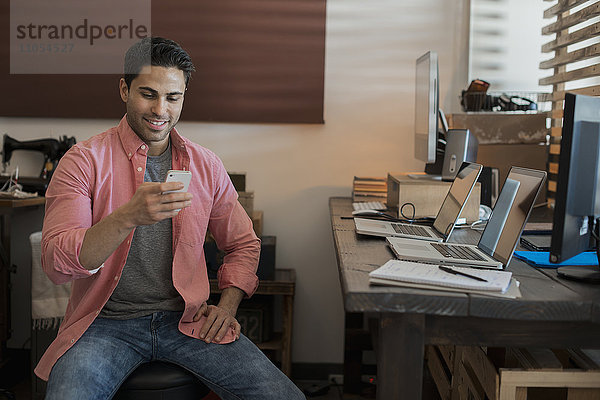 Ein Mann sitzt in einem Home-Office mit zwei Laptops auf dem Schreibtisch und überprüft sein Smartphone.