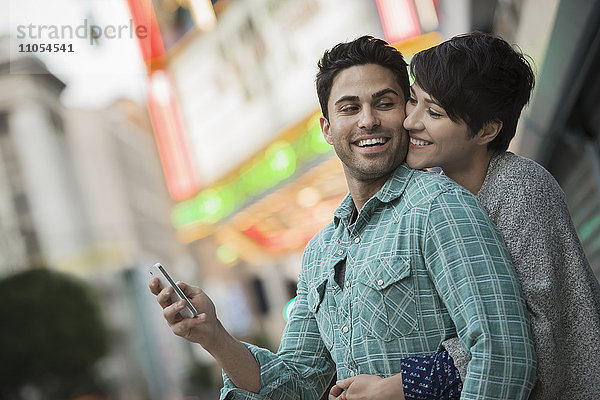 Ein Paar  Mann und Frau  die sich auf einer Straße der Stadt umarmen. Mann hält ein Smartphone in der Hand.