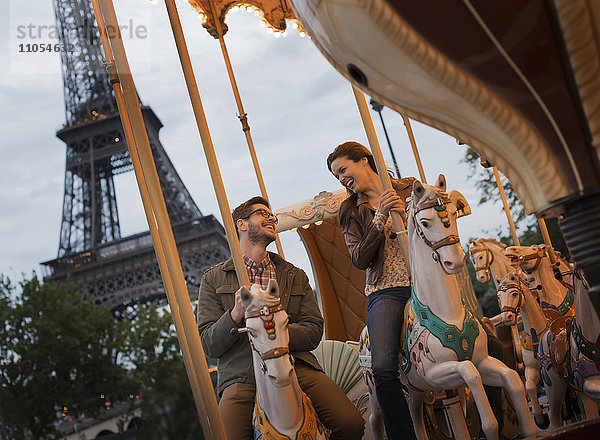 Ein Paar  Mann und Frau  reiten auf traditionellen Galoppern auf einem Karussell im Schatten des Eiffelturms in Paris.