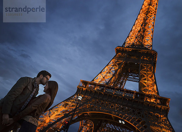 Ein Paar  das nachts auf dem Champs de Mars in Paris unter dem Windschatten des Eiffelturms steht.