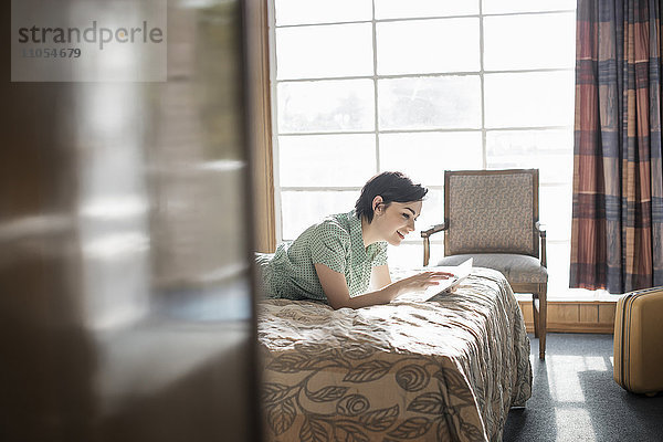 Eine junge Frau liegt auf einem Bett in einem Motelzimmer und benutzt einen digitalen Tablet-Touchscreen.