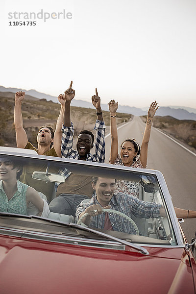 Eine Gruppe von Freunden in einem roten Cabrio-Oldtimer mit offenem Verdeck auf einer Autoreise.
