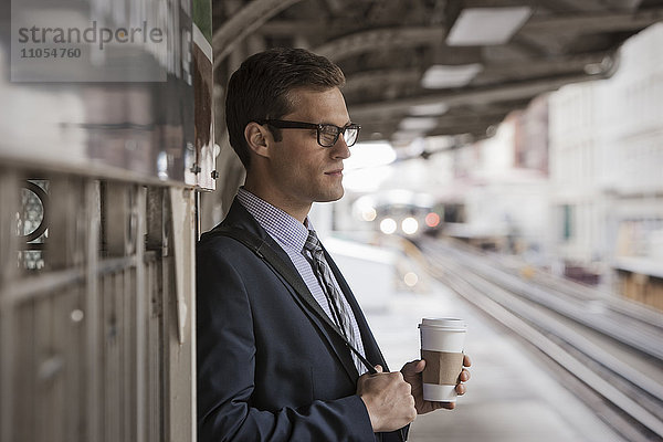 Ein Arbeitstag. Ein Geschäftsmann in Arbeitsanzug und Krawatte  der auf einem Bahnsteig eine Tasse Kaffee hält.
