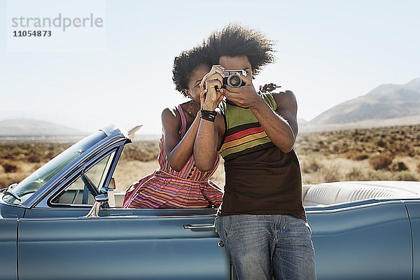 Ein junges Paar  Mann und Frau  an einem hellblauen Cabriolet auf offener Straße  mit einer Kamera in der Hand.