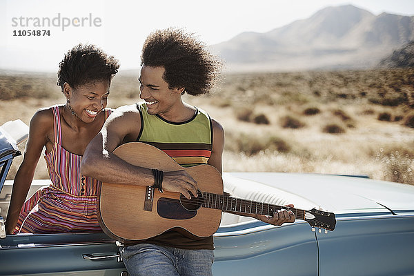 Ein junges Paar steht an einem hellblauen Cabriolet auf offener Straße  der Mann spielt Gitarre.