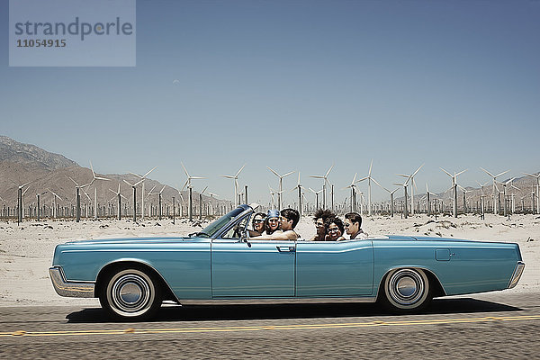 Eine Gruppe von Freunden in einem hellblauen Cabriolet auf offener Straße  die über eine trockene  flache  von Bergen umgebene Ebene fahren.