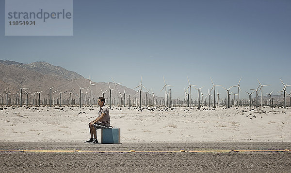 Ein Mann steht mit einem Koffer am Rande einer Autobahn  mit Windturbinen im Hintergrund.
