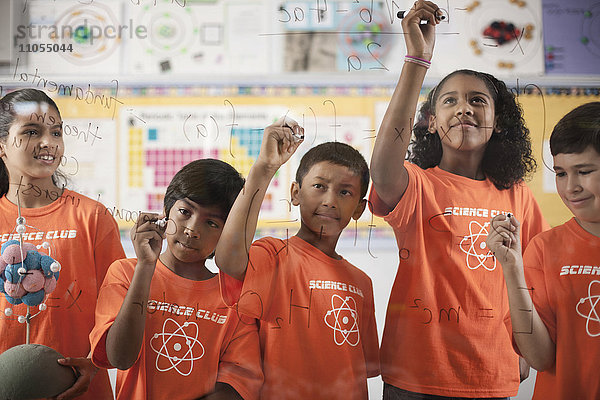 Eine Gruppe von Mädchen und Jungen  die das T-Shirt des Science Club tragen und Gleichungen und Formeln auf eine durchsichtige Tafel schreiben.