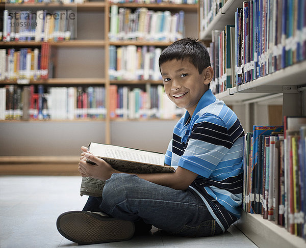 Ein Junge sitzt in einer Bibliothek auf dem Boden und liest ein Buch.