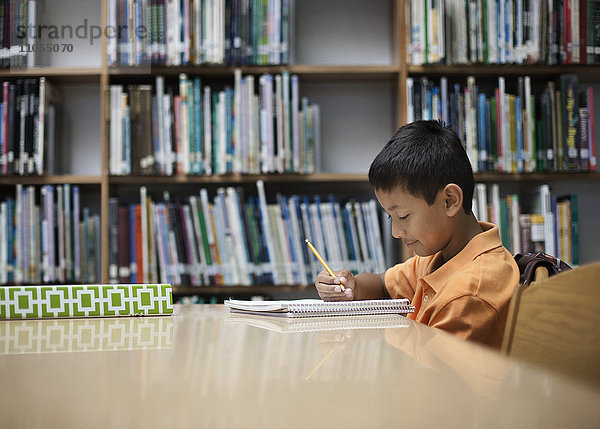 Ein Junge  der in einer Schulbibliothek am Tisch sitzt und mit einem Bleistift lernt.
