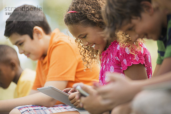 Eine Reihe von Kindern  die im Sommer mit Tabletten und Handspielen im Freien sitzen.