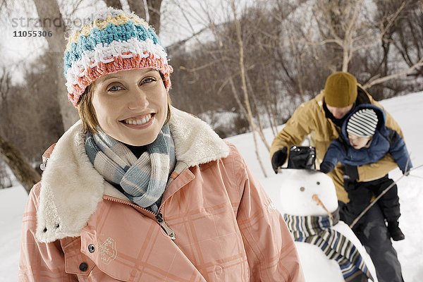 Eine Frau und ein Mann halten ein Kind und inspizieren einen Schneemann mit Hut und Schal.