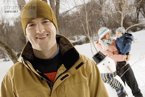 Im Vordergrund ein lächelnder Mann und eine Frau  die ein Kind neben einem Schneemann hält.