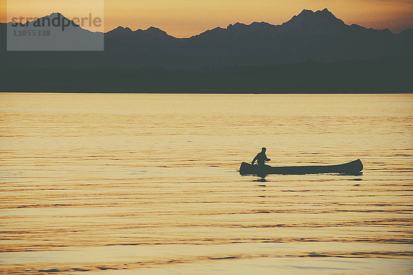 Eine Person sitzt in einem großen Kanu im Indianerstil und paddelt bei Sonnenuntergang über ruhiges Wasser.