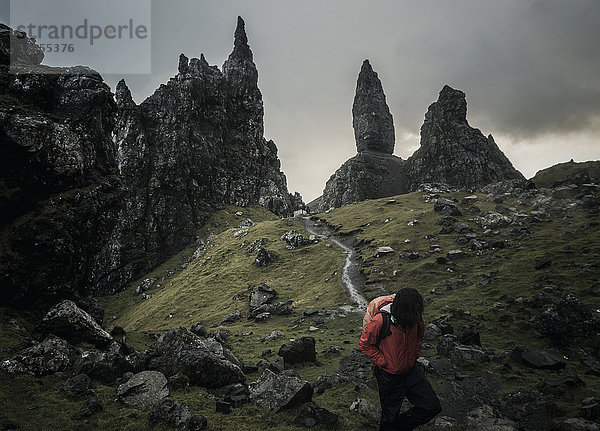 Zwei Personen mit Rucksäcken auf einem schmalen Pfad  der zu einer dramatischen Landschaft aus Felsspitzen in der sie überragenden Skyline führt  unter einem bedeckten Himmel mit niedriger Bewölkung.