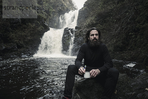 Wildes Camping. Ein Mann sitzt mit einem Becher in der Hand  an einem schnell fließenden Bach und kaskadenartigem Wasser.
