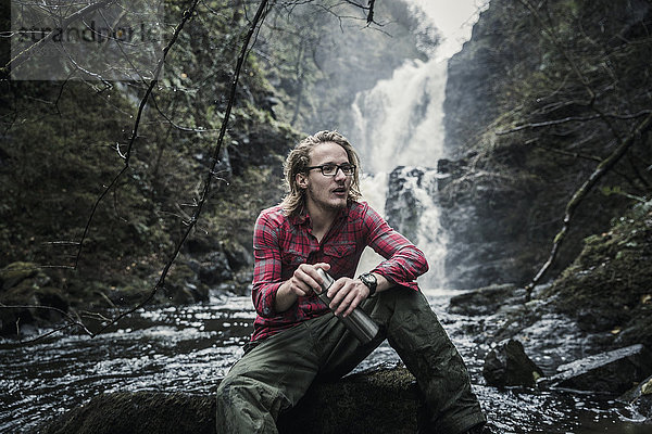 Ein Mann sitzt auf einem Felsen bei einem Wasserfall und hält einen Flachmann. Winterwandern.