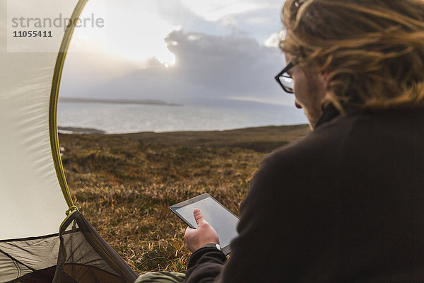 Ein Mann sitzt im Schutz eines Zeltes  schaut hinaus und hält ein digitales Tablet