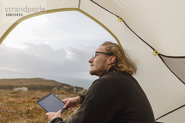 Ein Mann sitzt im Schutz eines Zeltes  schaut hinaus und hält ein digitales Tablett in der Hand.