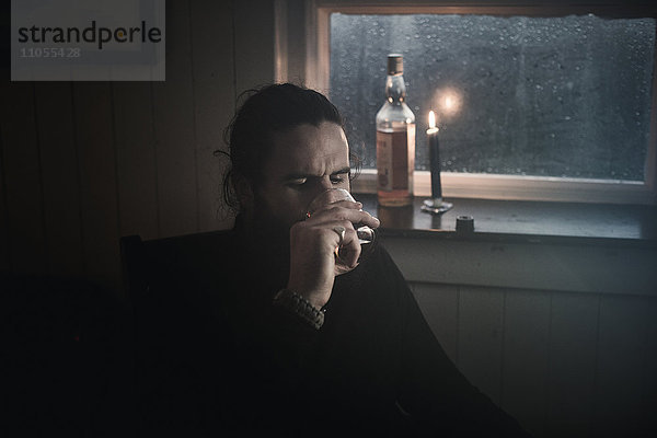 Ein Mann sitzt im Dunkeln an einem Fenster bei Kerzenlicht und trinkt aus einem kleinen Glas. Eine Flasche neben ihm.