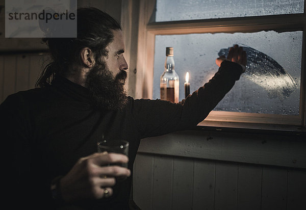 Ein Mann sitzt allein in einem Raum mit einer Flasche Whisky und einem Glas und wischt Kondenswasser vom Fenster  um nach draußen zu sehen. Eine brennende Kerze.