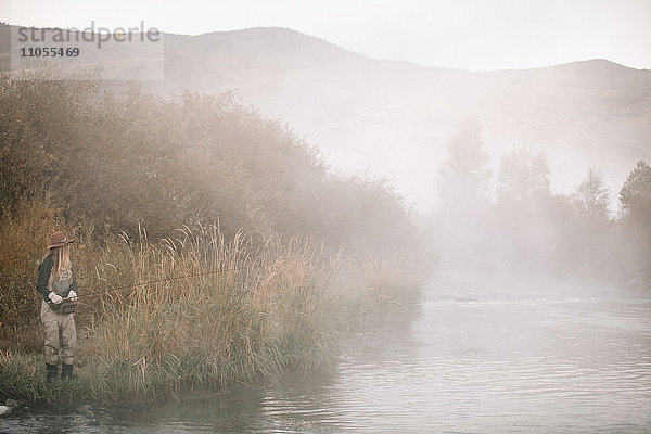 Eine Frau beim Angeln  am Flussufer stehend. Nebel steigt aus dem Wasser auf.