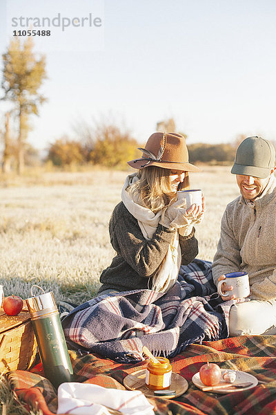 Zwei Personen  ein Paar bei einem Winterpicknick  sitzen auf einem Schottenkaro-Teppich und halten heiße Getränke in der Hand.